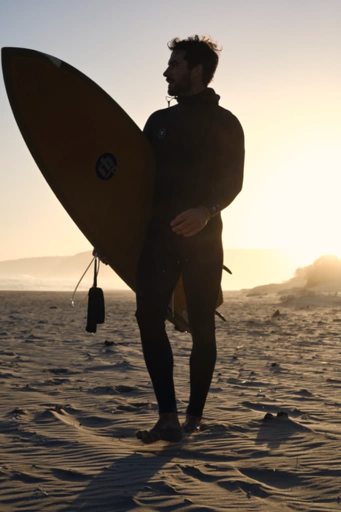 Marc Charbonneau in a wetsuit with a surfboard on the beach at golden hour.